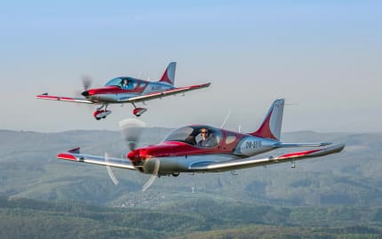 Bristell Classic and Bristell RG flying in formation over wooded hills, the RG with its gear retracted