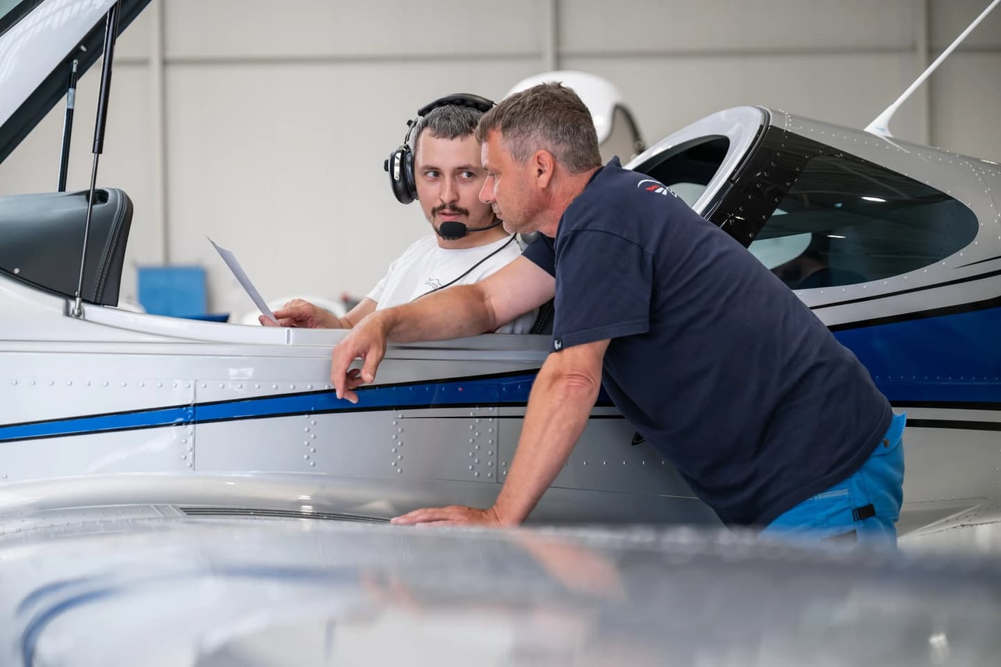 Two men going through a printed document at a Bristell aircraft in the hangar, one seated in the open cockpit wearing an aviation headset