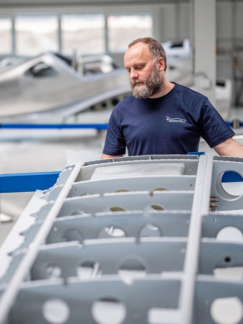 A Bristell technician beside a wing structure in the assembly hall, its primed ribs and lightening holes still open