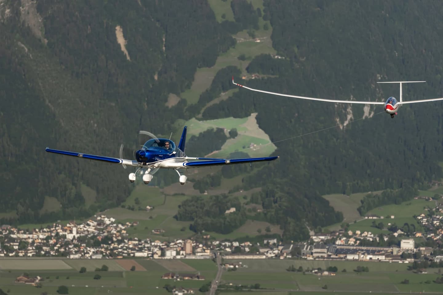 A blue Bristell aircraft towing a glider over an alpine valley