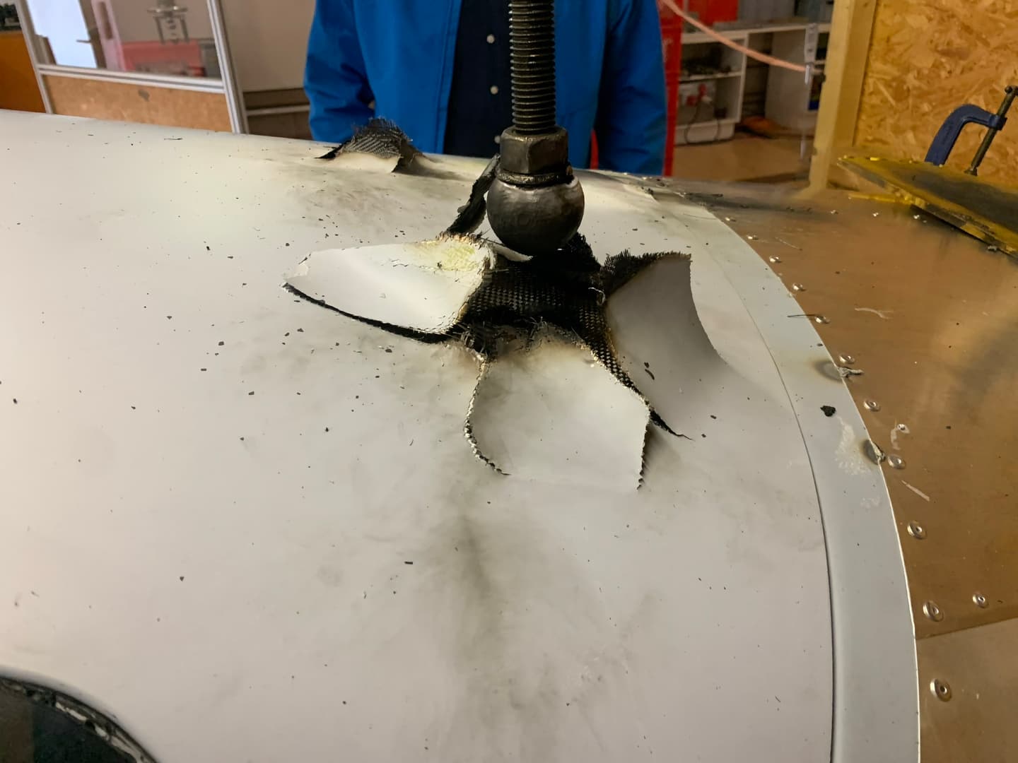 A Bristell airframe section on a laboratory lightning-strike test rig — the discharge electrode pressed to the surface, the direct-strike contact point scorched and torn open
