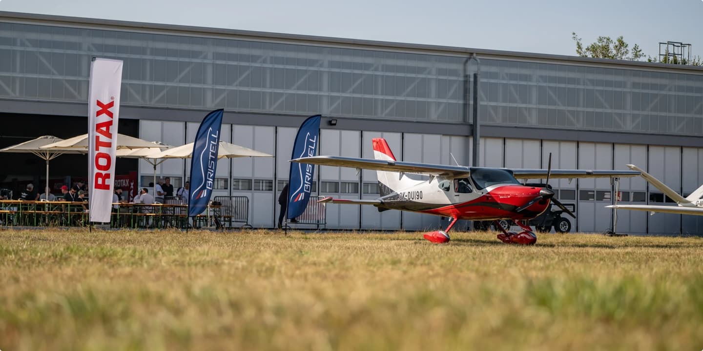 A Bristell aircraft on display at an aviation event, with Bristell and Rotax branded flags in the background