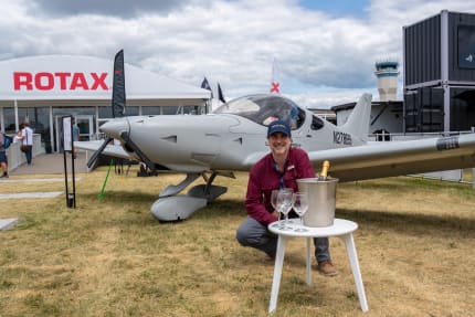 Bernardo Saviñon crouching beside his military-gray Bristell Classic N278BS with a champagne bucket at the Rotax exhibit, EAA AirVenture Oshkosh 2026