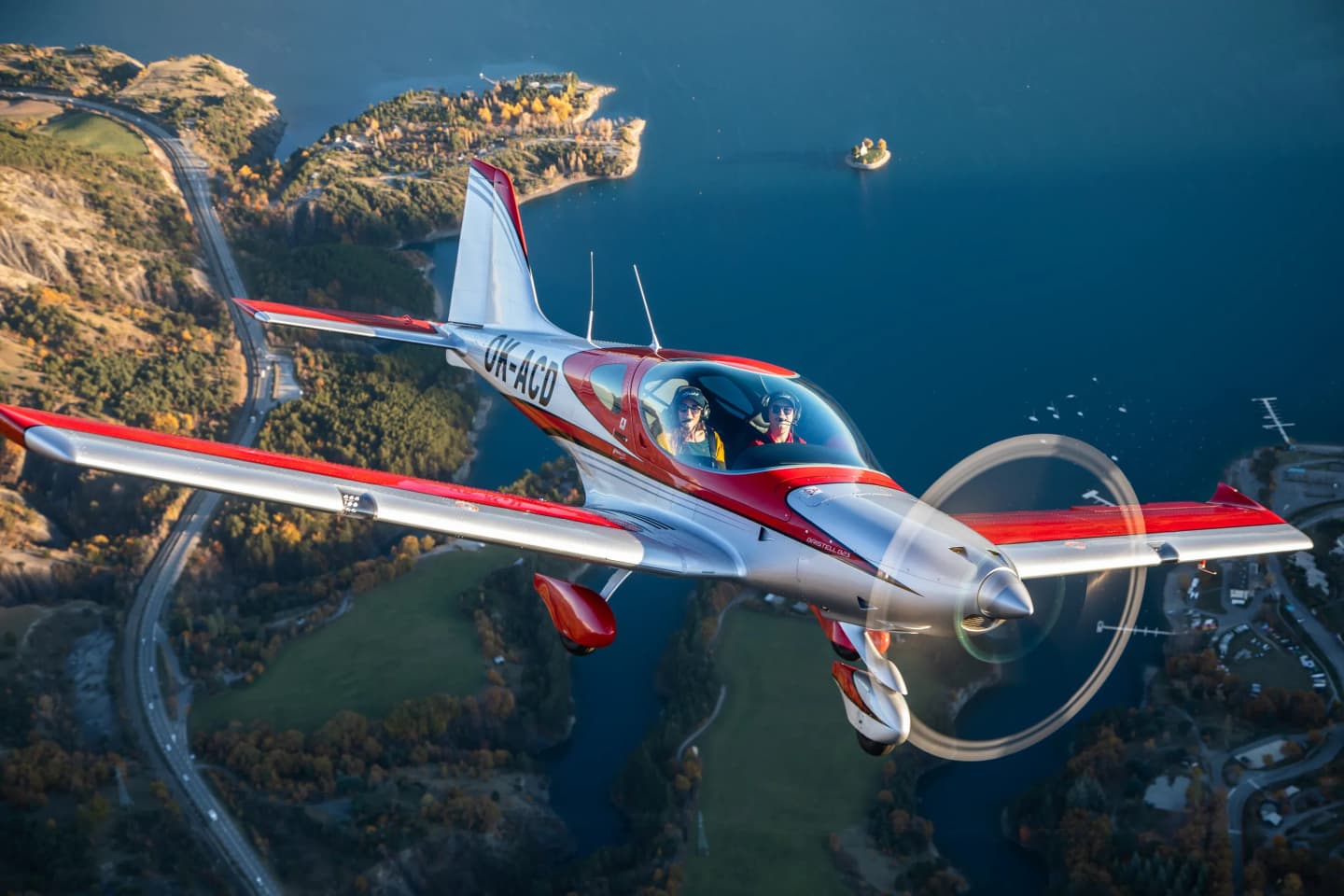 Red and silver Bristell B23 with two aboard flying over a lake and autumn shoreline