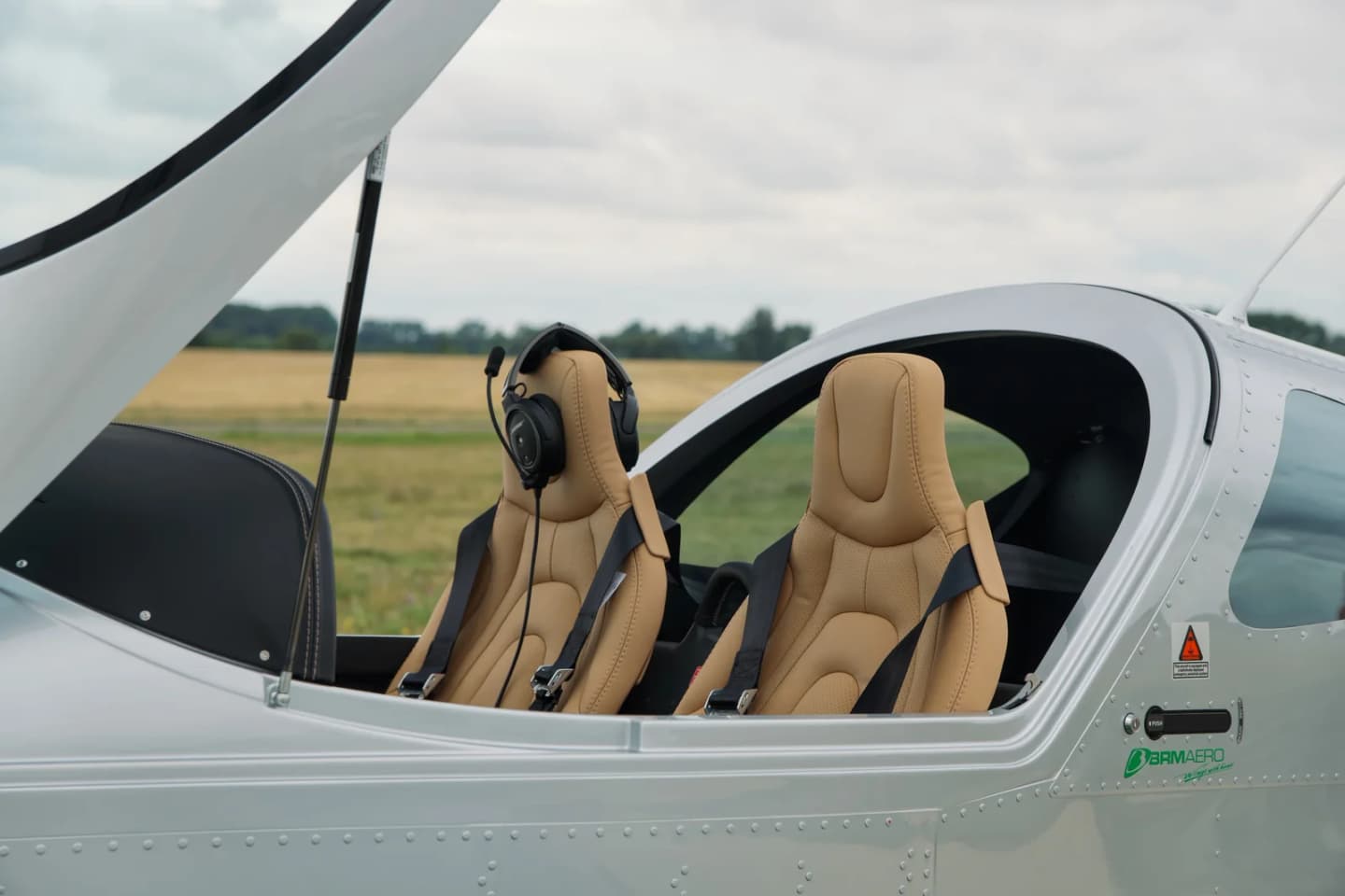 Two tan perforated leather seats side by side under the open canopy of the Bristell B23, with a headset resting on the left seat