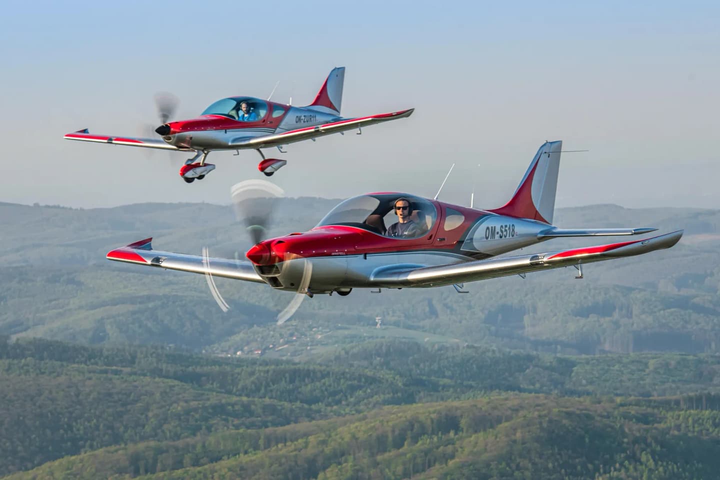 Two Bristell light aircraft in red and polished-metal livery flying in formation over forested hills
