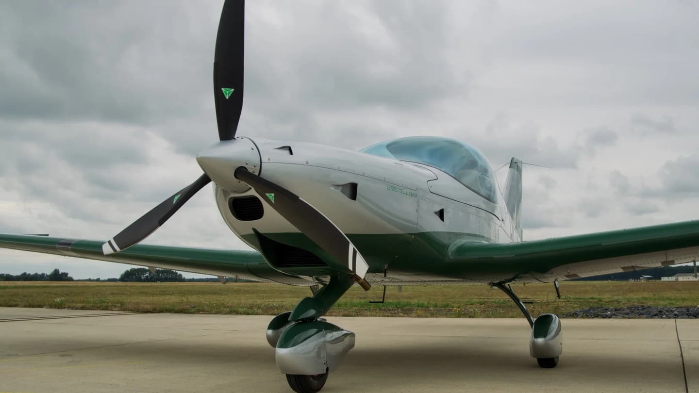 Factory-new Bristell B23 in silver and green livery parked on the apron, seen from the front three-quarter angle with its three-blade constant-speed propeller