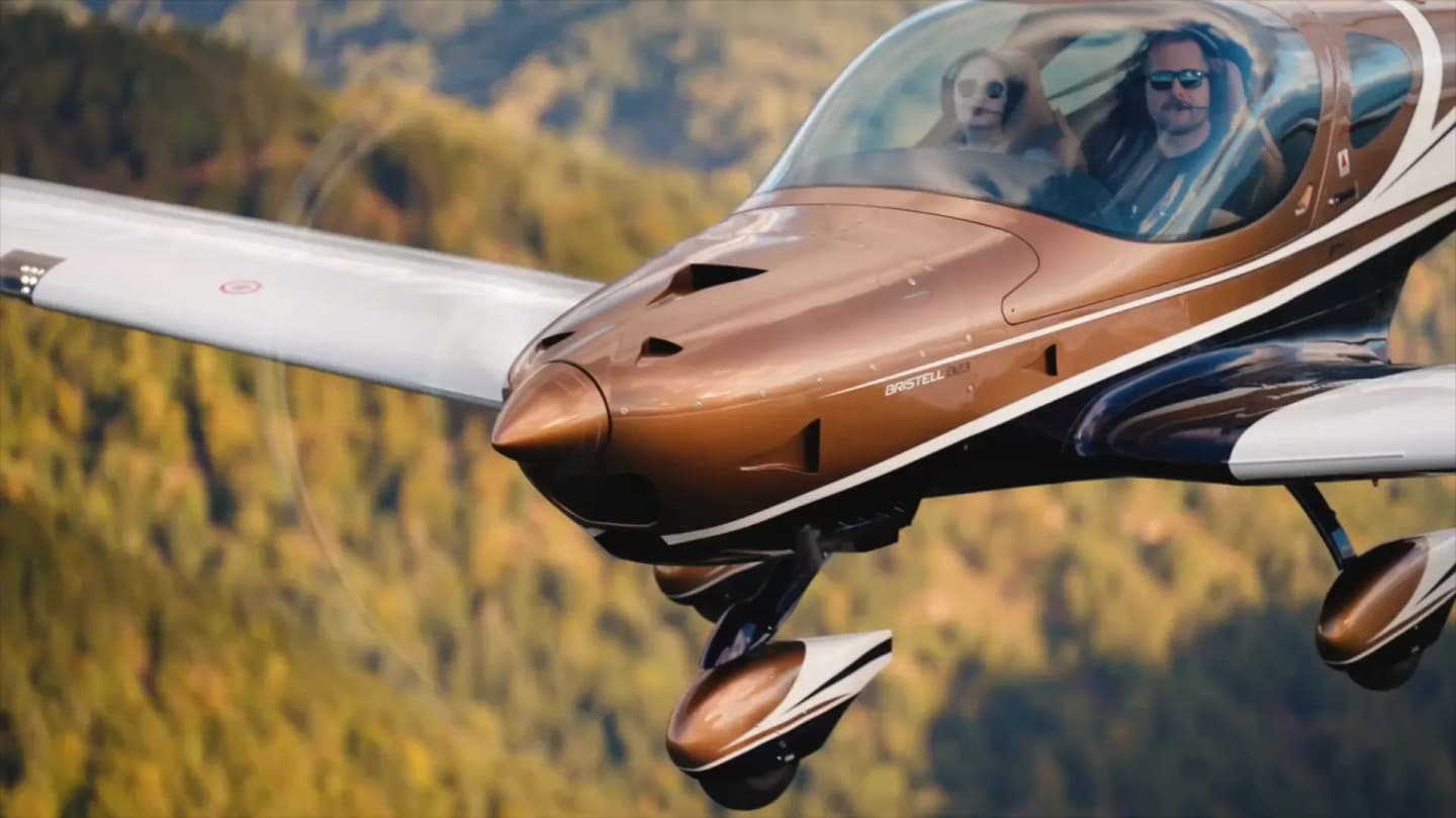 Bronze-and-white Bristell B23 with pilot and passenger flying low over autumn forested hills