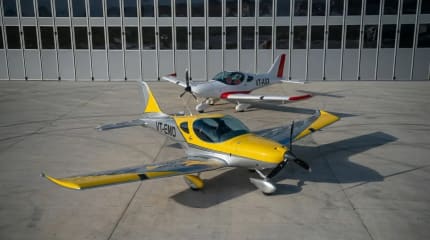 Two Bristell aircraft parked on the apron in front of a hangar, one in yellow and silver in the foreground and one in white and red behind it