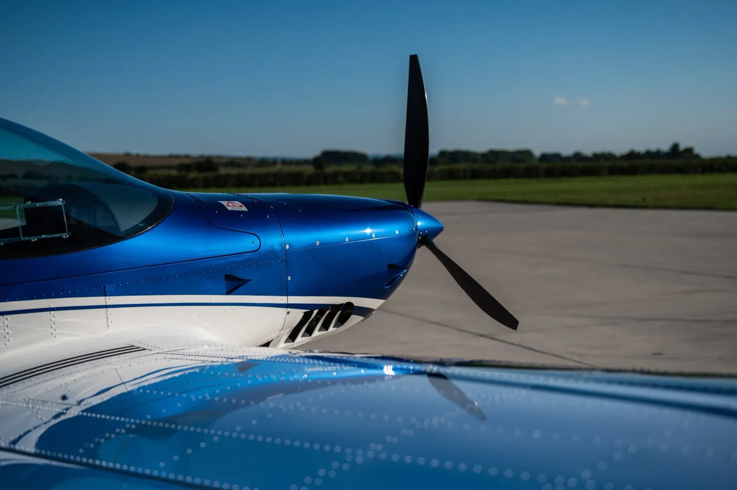 Side view of the blue-and-white nose, spinner and black propeller of a Bristell aircraft parked on an airfield apron, with green fields behind.