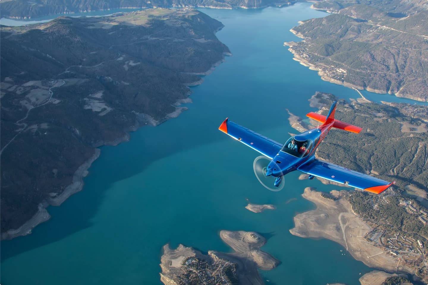 A two-seat Bristell aircraft in flight over a lake and mountain landscape, with pilot and passenger visible in the cockpit